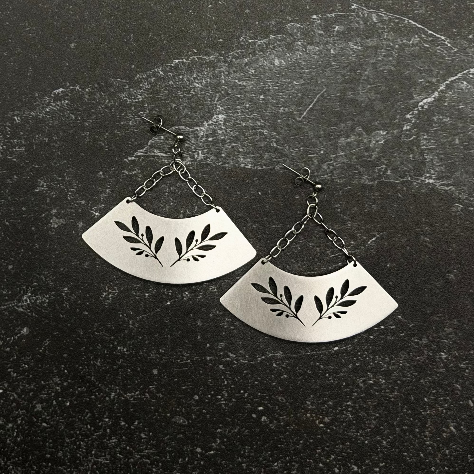 Stainless steel fan-shaped earrings with leaf design on a dark textured surface