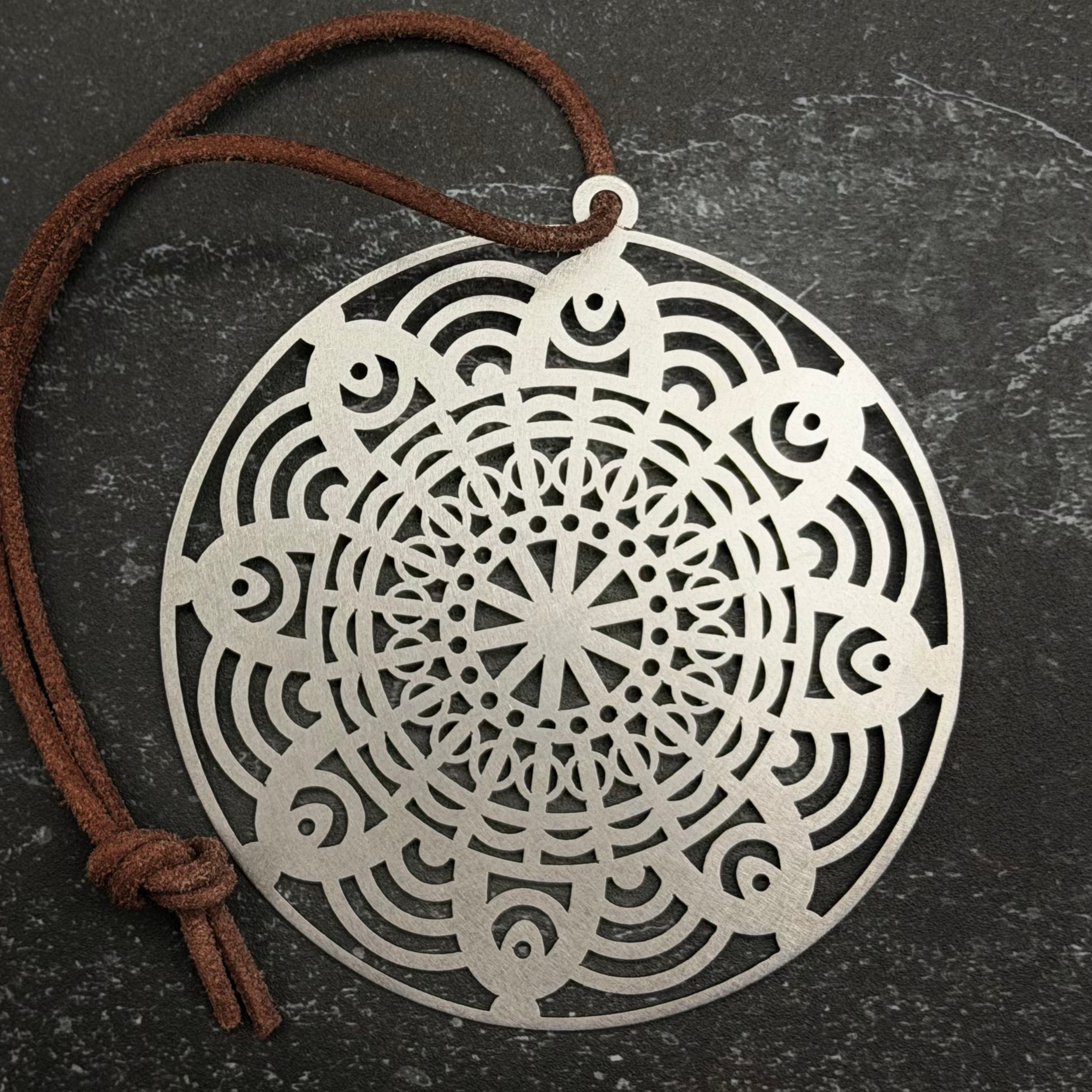 Stainless steel circular metal pendant with intricate patterns on a dark surface