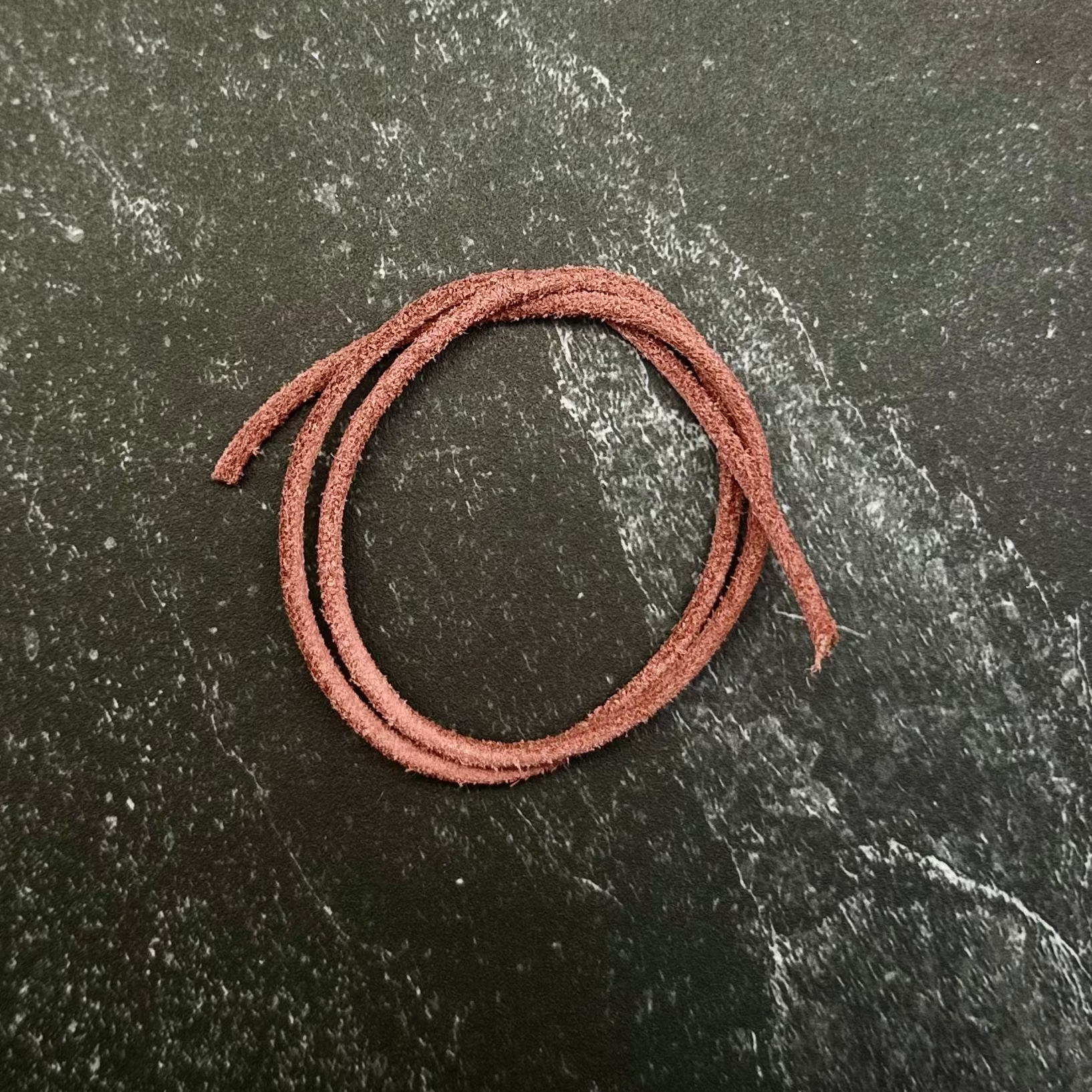 A small coiled piece of reddish-brown cord or thread placed on a dark hexagonal stone slab, set against a dark black fabric background.