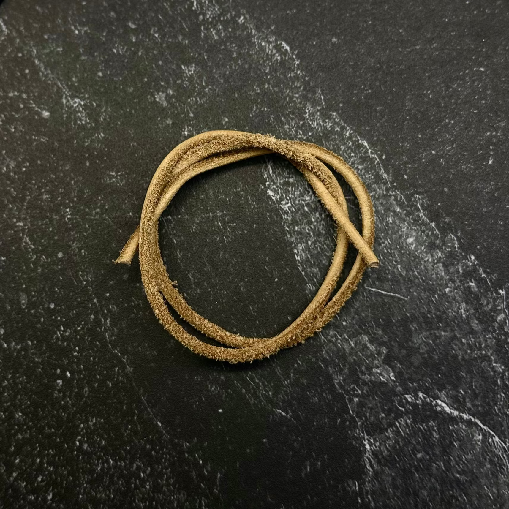 A gold-colored braided cord or strap, coiled in a circle, displayed on a dark gray hexagonal stone slab set against a dark navy blue fabric background.