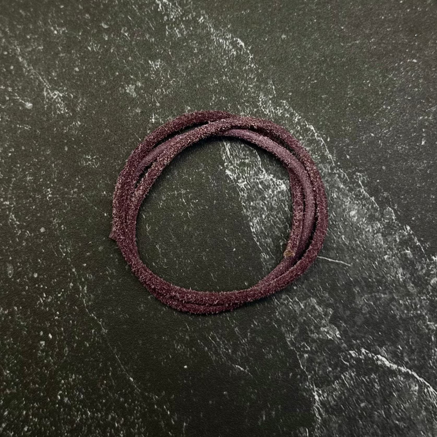 A dark burgundy braided hair tie or elastic band displayed on a hexagonal dark stone slab, set against a dark black fabric background.