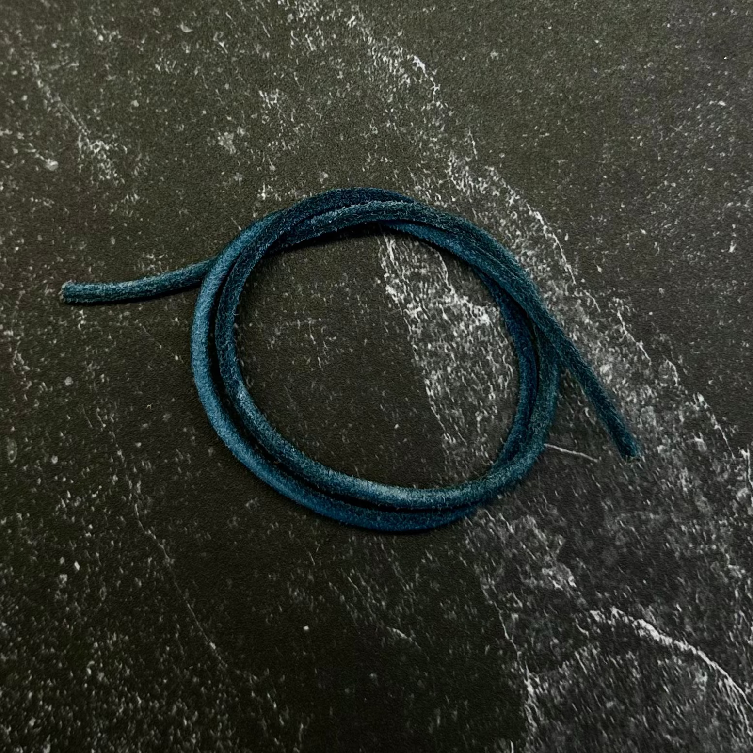 A dark teal blue cord or strap coiled on a hexagonal dark stone slab, placed on top of a black fabric background.