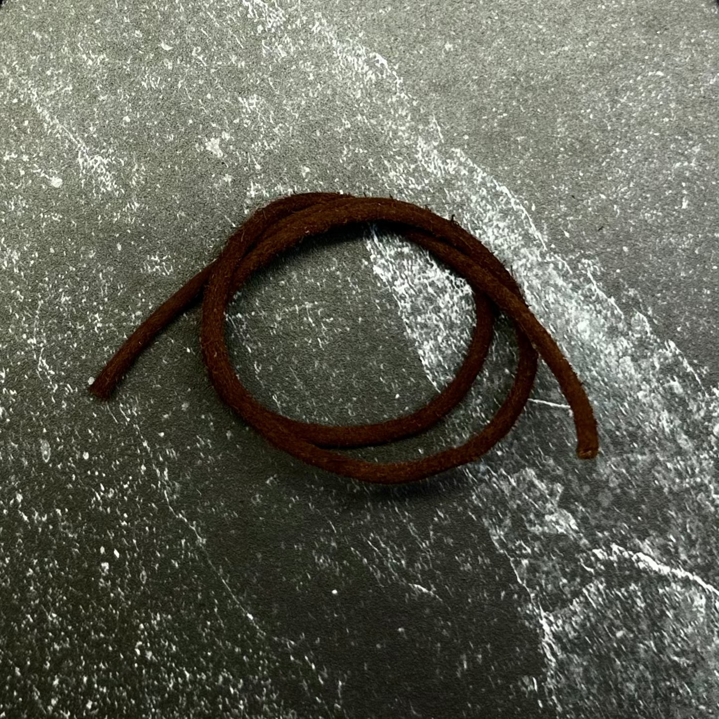 A dark brown braided cord or strap coiled on a hexagonal gray stone slab, set against a dark charcoal gray fabric background.