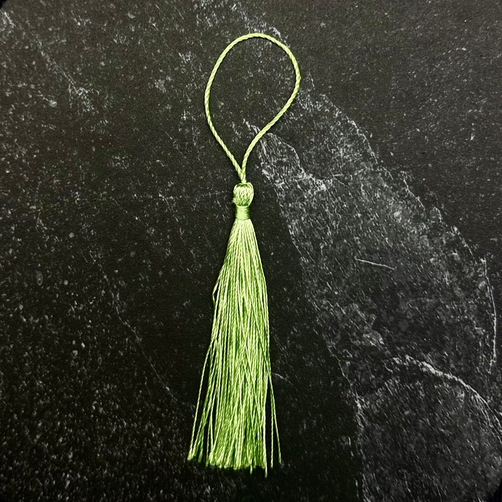 A light green tassel ornament with a thin loop at the top, displayed on a dark hexagonal stone tile against a black fabric background.