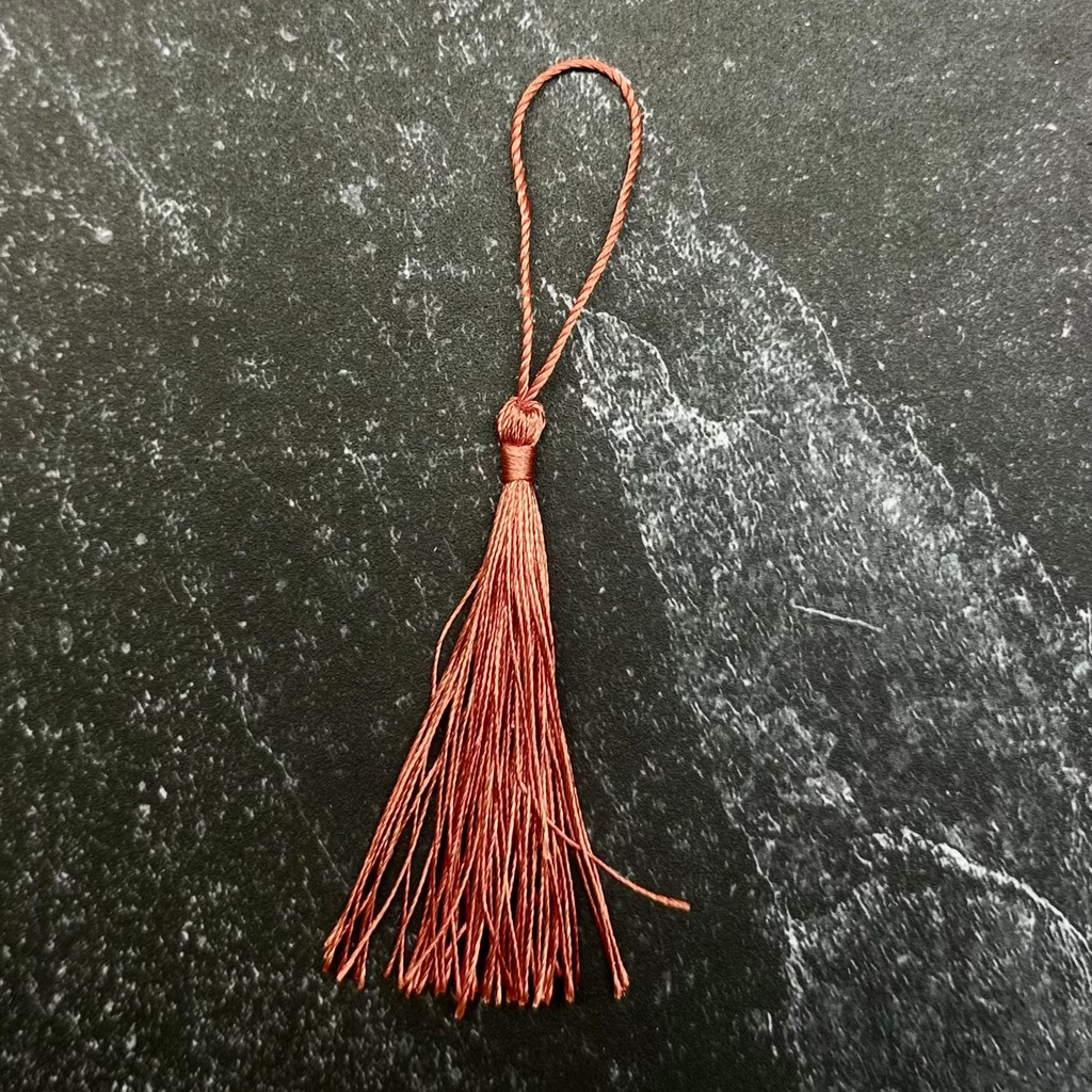 A rust-brown leather tassel with a loop at the top, displayed on a dark green hexagonal stone tile over a black ribbed fabric background.
