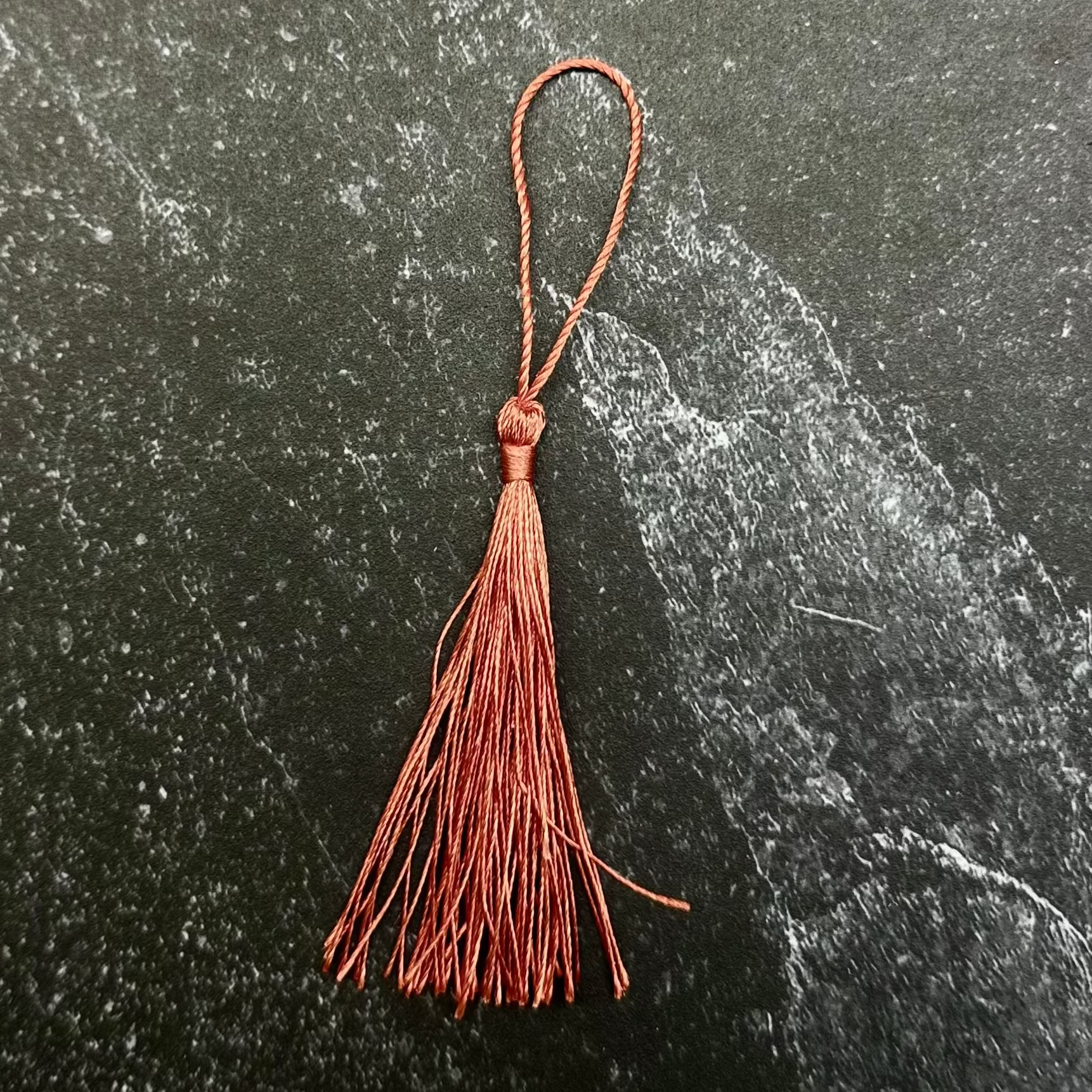 A rust-brown leather tassel with a loop at the top, displayed on a dark green hexagonal stone tile over a black ribbed fabric background.