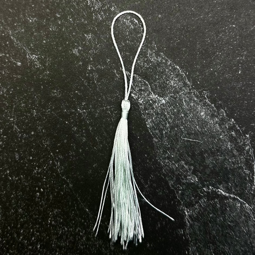 A small white tassel ornament with a silver metal loop, displayed on a dark hexagonal stone slab against a black fabric background.