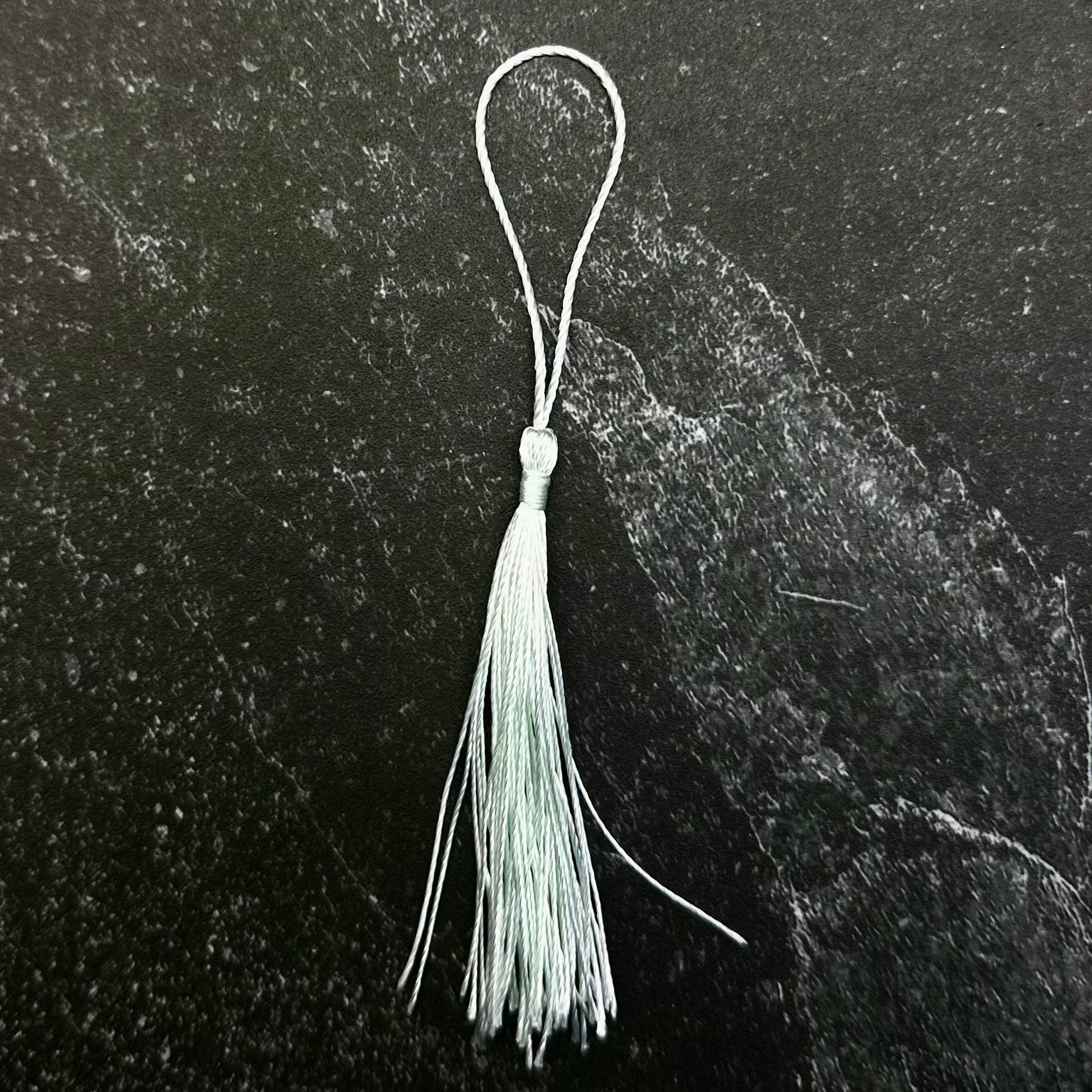 A small white tassel ornament with a silver metal loop, displayed on a dark hexagonal stone slab against a black fabric background.