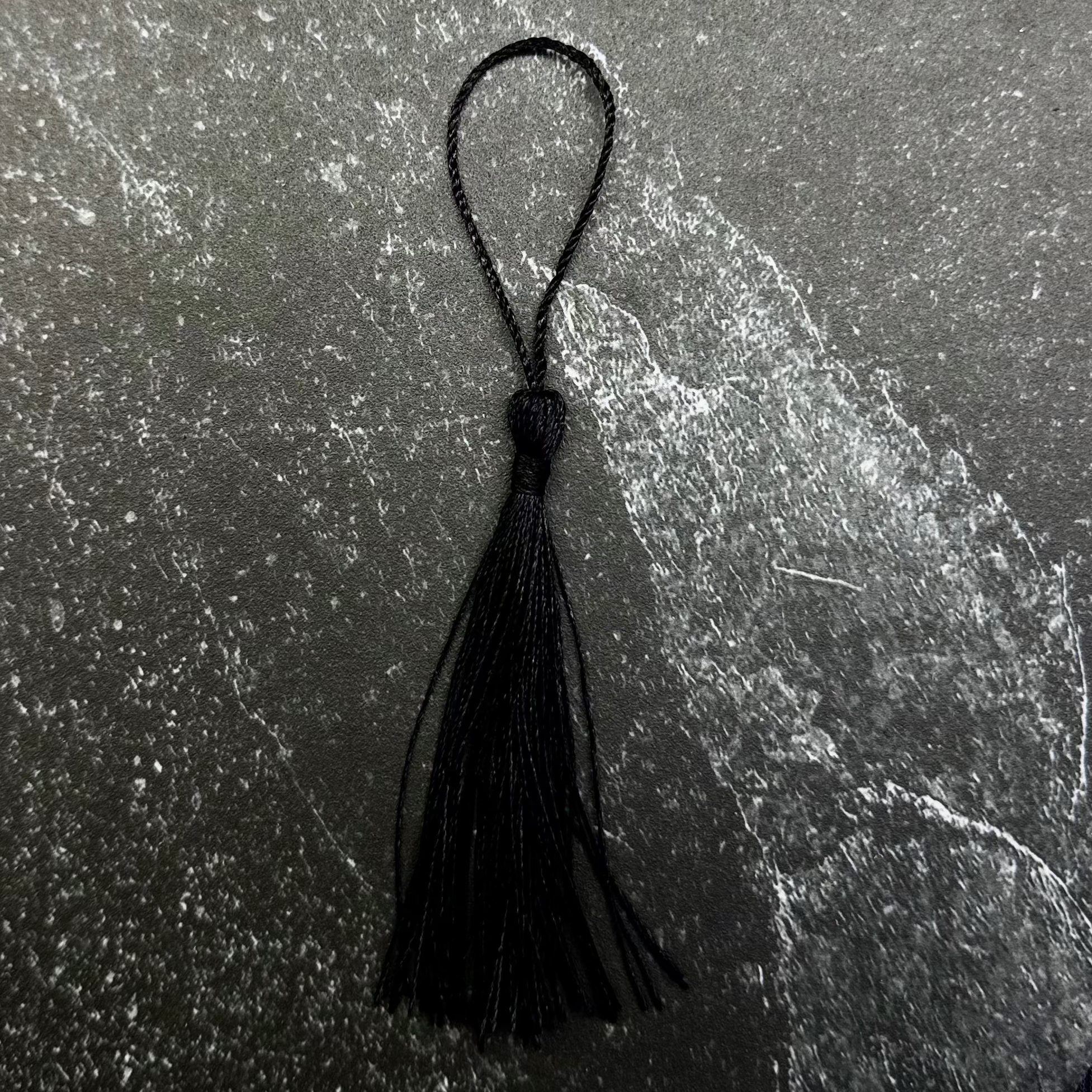 A black tassel with a matching black loop, displayed on a dark gray hexagonal stone tile over a navy blue ribbed fabric background.