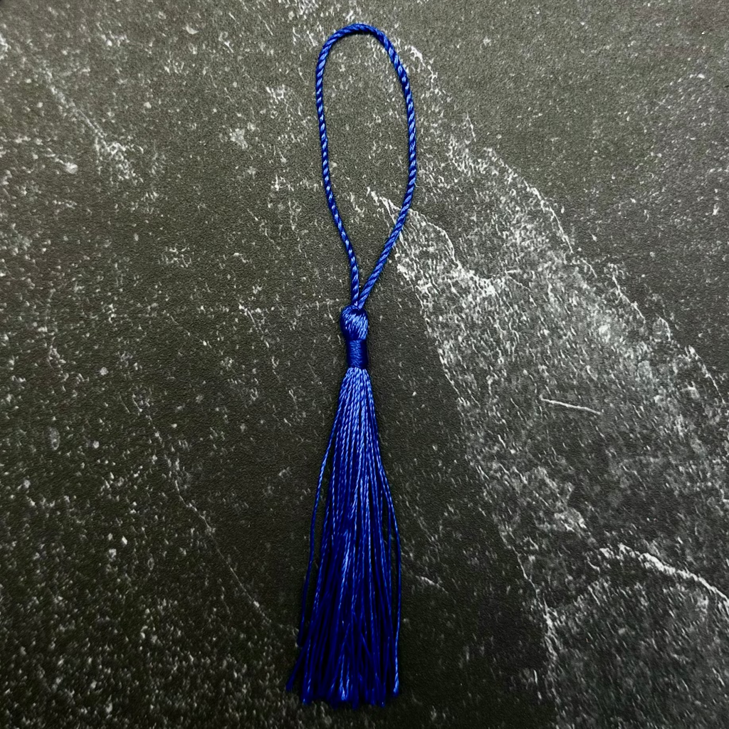 A royal blue tassel with a matching blue loop, displayed on a dark gray hexagonal stone slab set against a black ribbed fabric background.