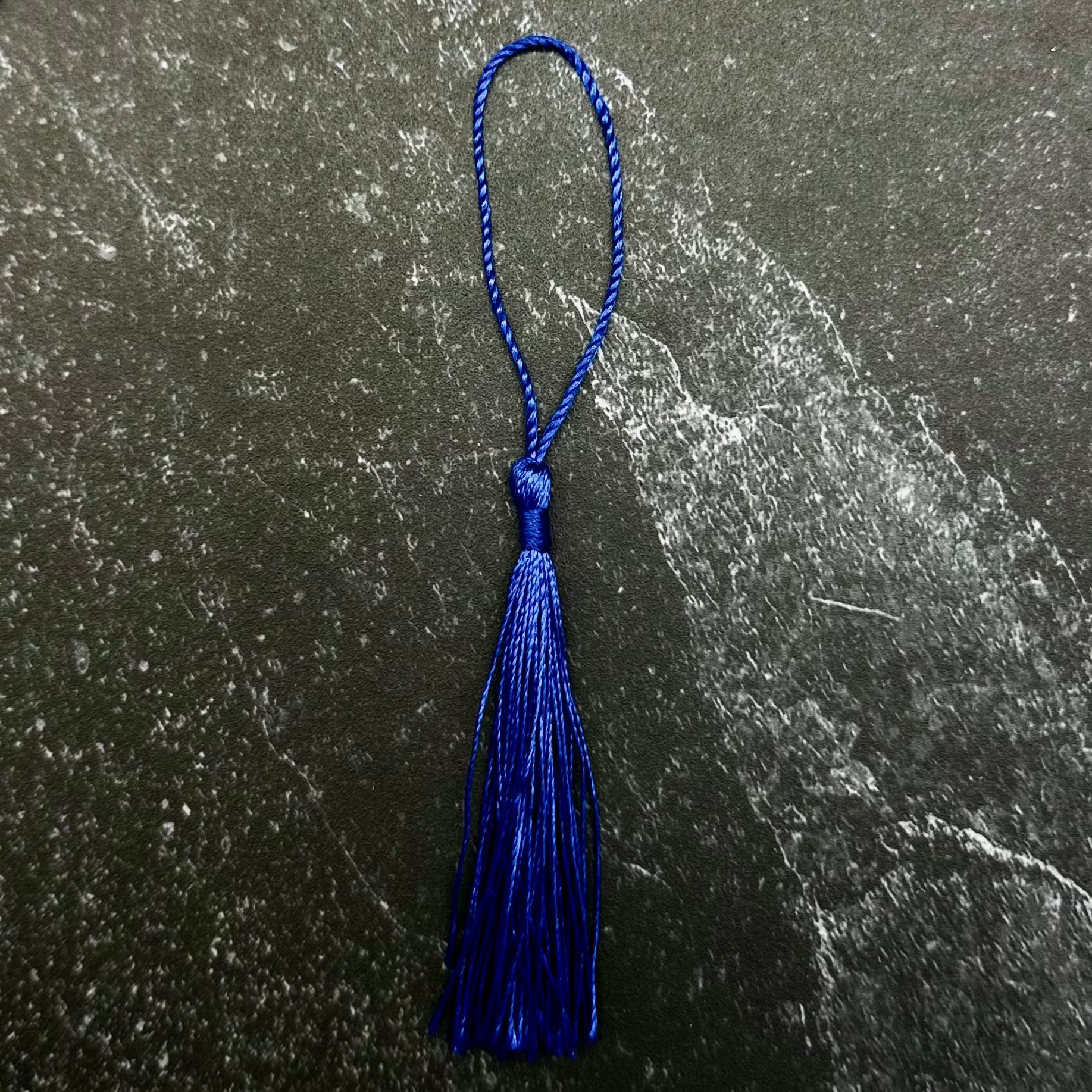 A royal blue tassel with a matching blue loop, displayed on a dark gray hexagonal stone slab set against a black ribbed fabric background.