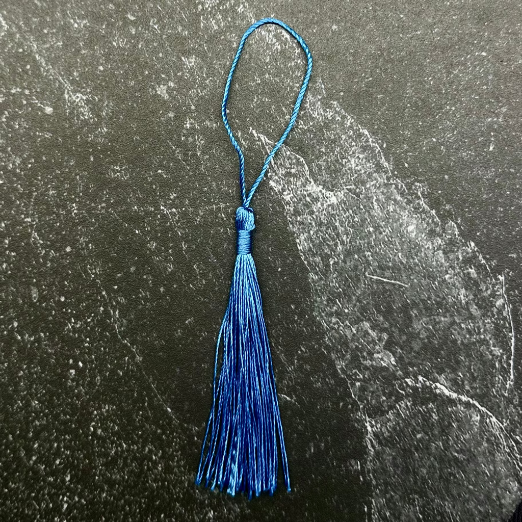 A small blue tassel ornament with a loop at the top, placed on a dark hexagonal stone slab against a black ribbed fabric background.