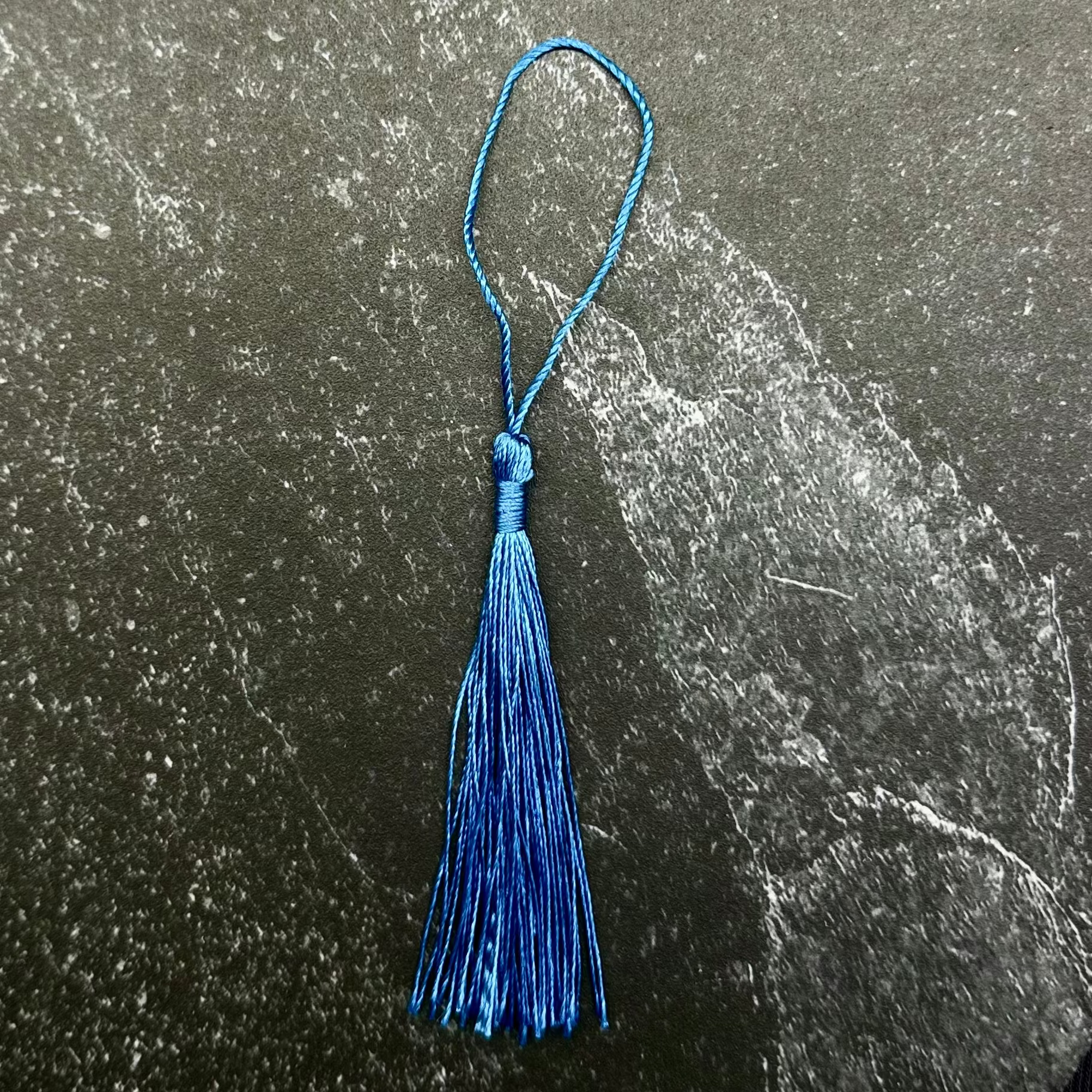 A small blue tassel ornament with a loop at the top, placed on a dark hexagonal stone slab against a black ribbed fabric background.