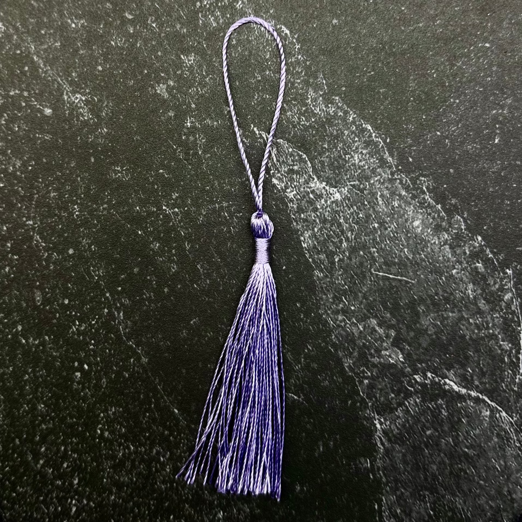 A small purple tassel ornament with a matching loop at the top, displayed on a dark green hexagonal stone slab against a black fabric background.