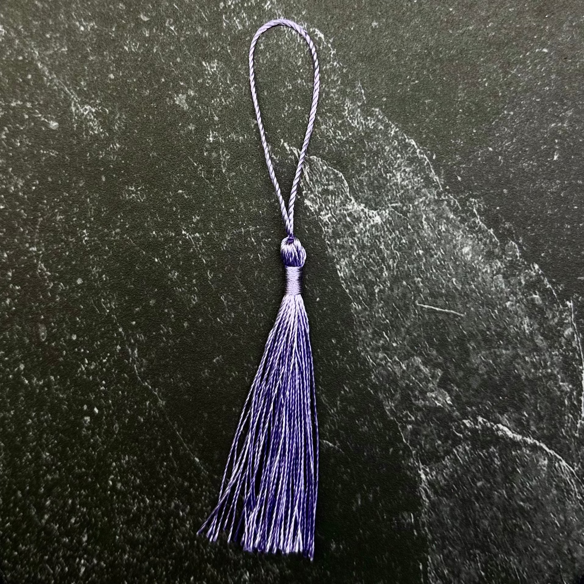 A small purple tassel ornament with a matching loop at the top, displayed on a dark green hexagonal stone slab against a black fabric background.