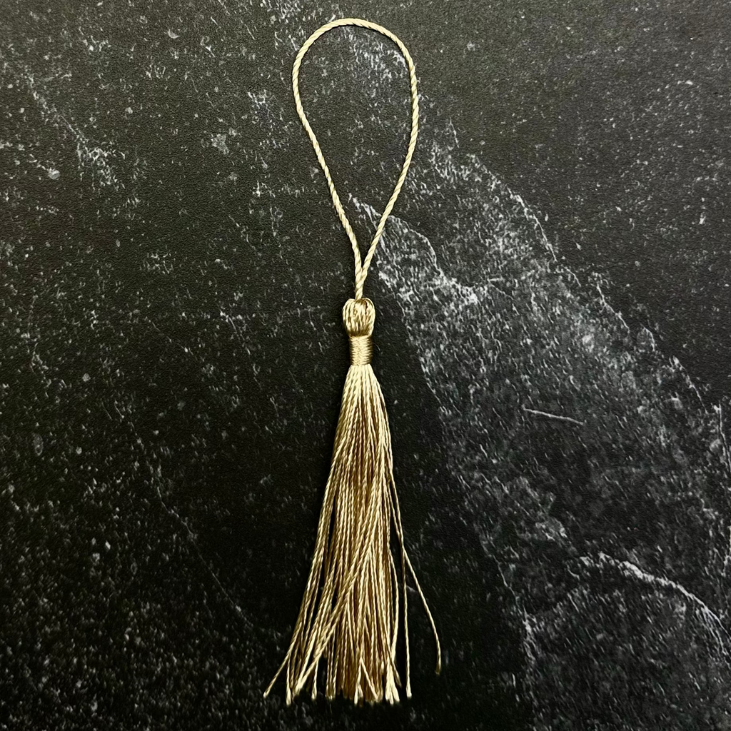 A small gold tassel ornament with a thin hanging loop, displayed on a dark hexagonal stone slab against a black fabric background.