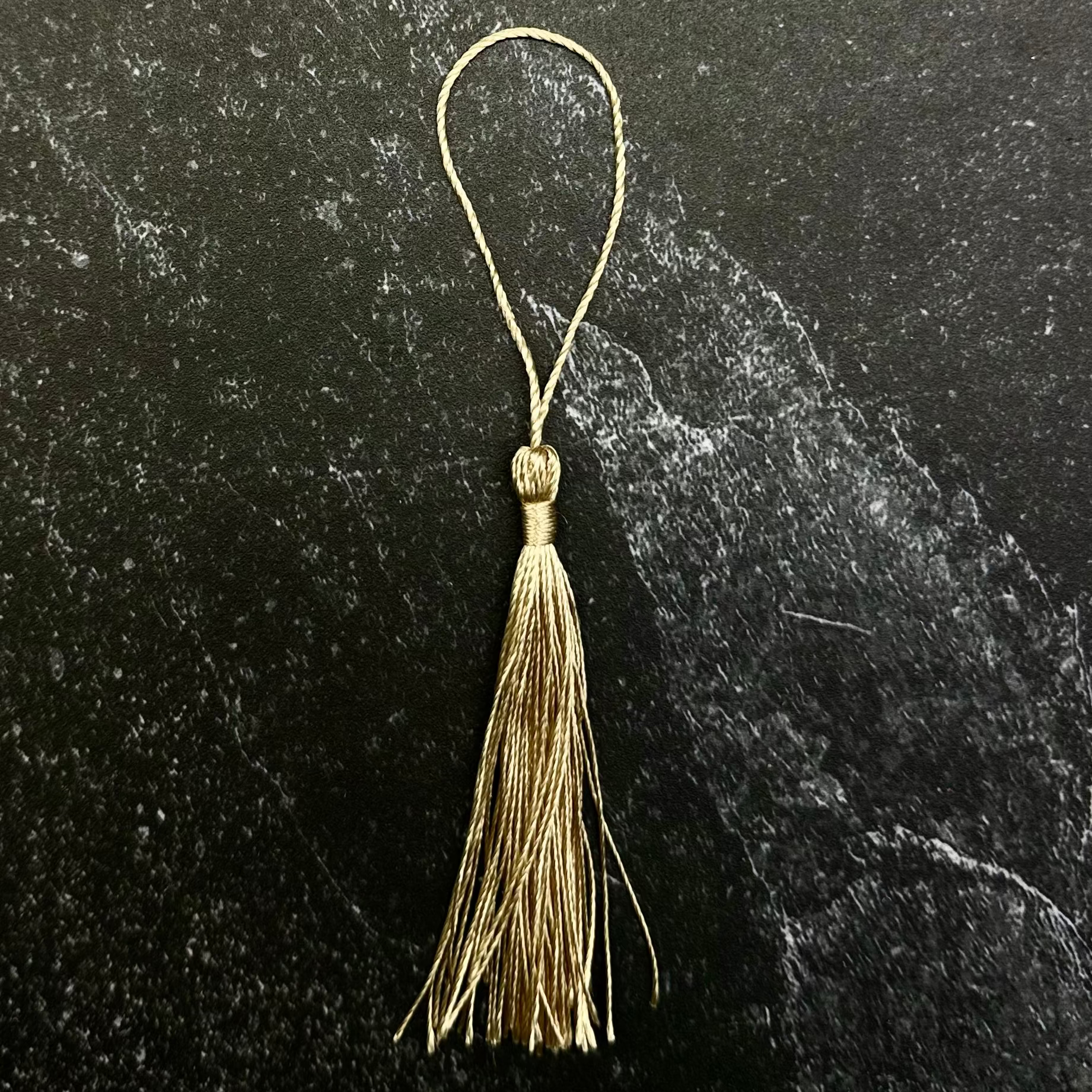A small gold tassel ornament with a thin hanging loop, displayed on a dark hexagonal stone slab against a black fabric background.
