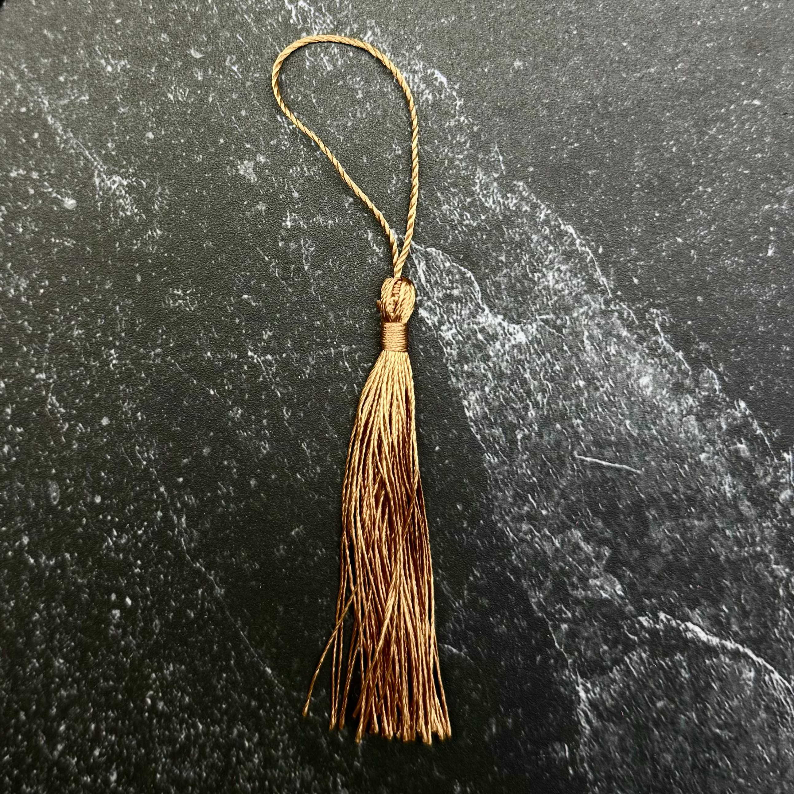 A small tan tassel ornament with a loop at the top, resting on a dark hexagonal stone slab against a black fabric background.