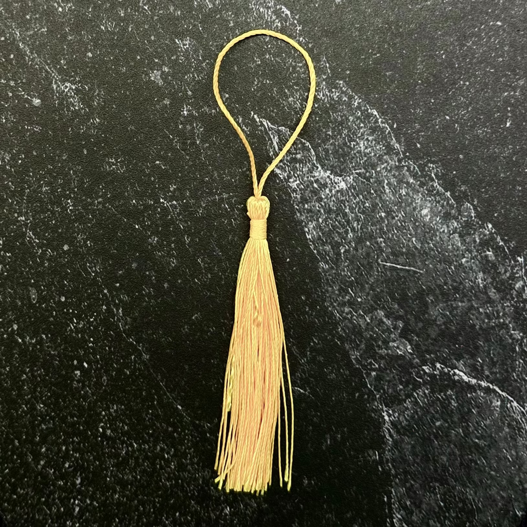 A gold tassel ornament with a matching gold loop, displayed on a dark hexagonal slate tile over a black fabric background.