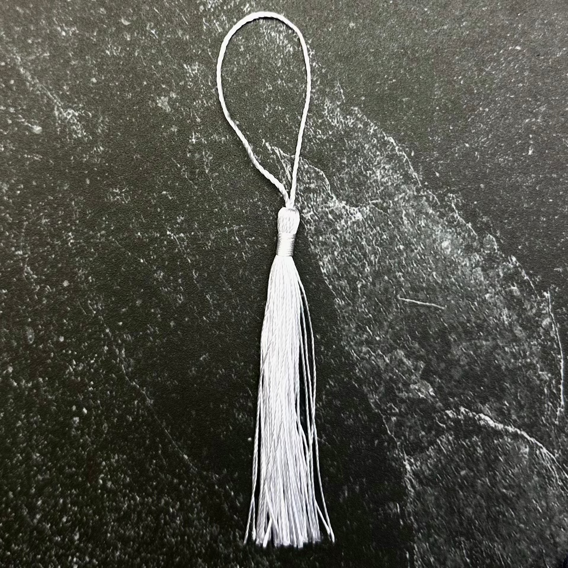 A white tassel with a thin white loop string, displayed on a dark gray hexagonal stone plate over a black ribbed fabric background.