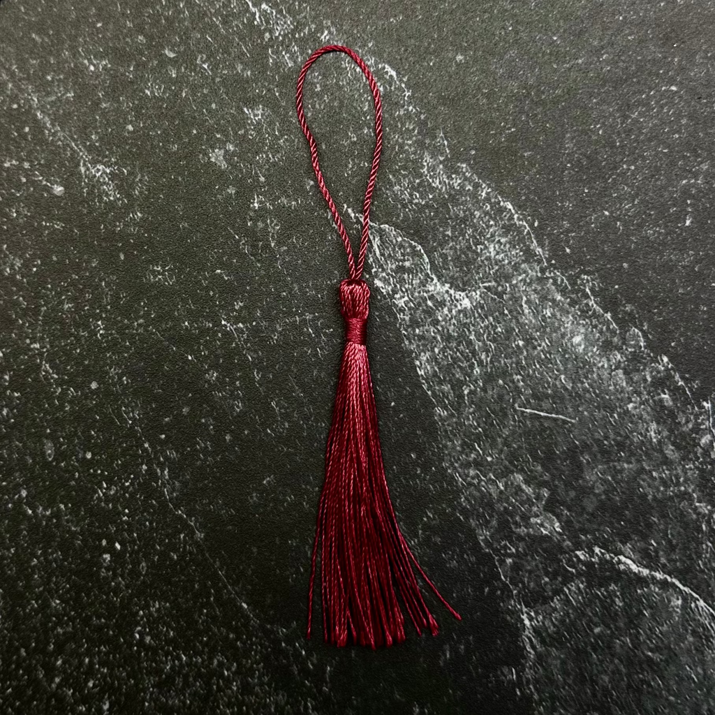 A dark red tassel with a matching loop, displayed on a hexagonal dark stone tile against a black ribbed fabric background.