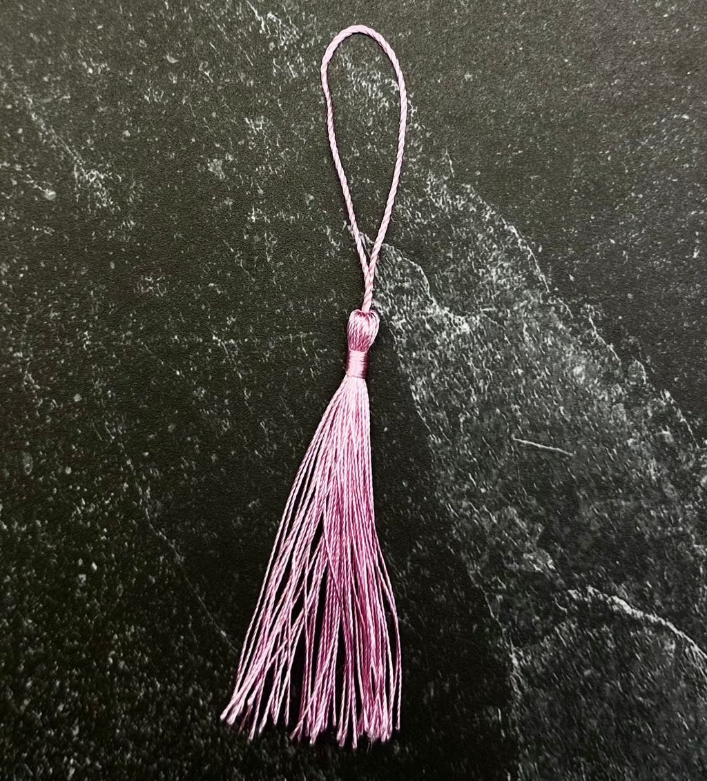 A pink tassel ornament with a matching pink loop, displayed on a dark hexagonal stone tile against a black fabric background.