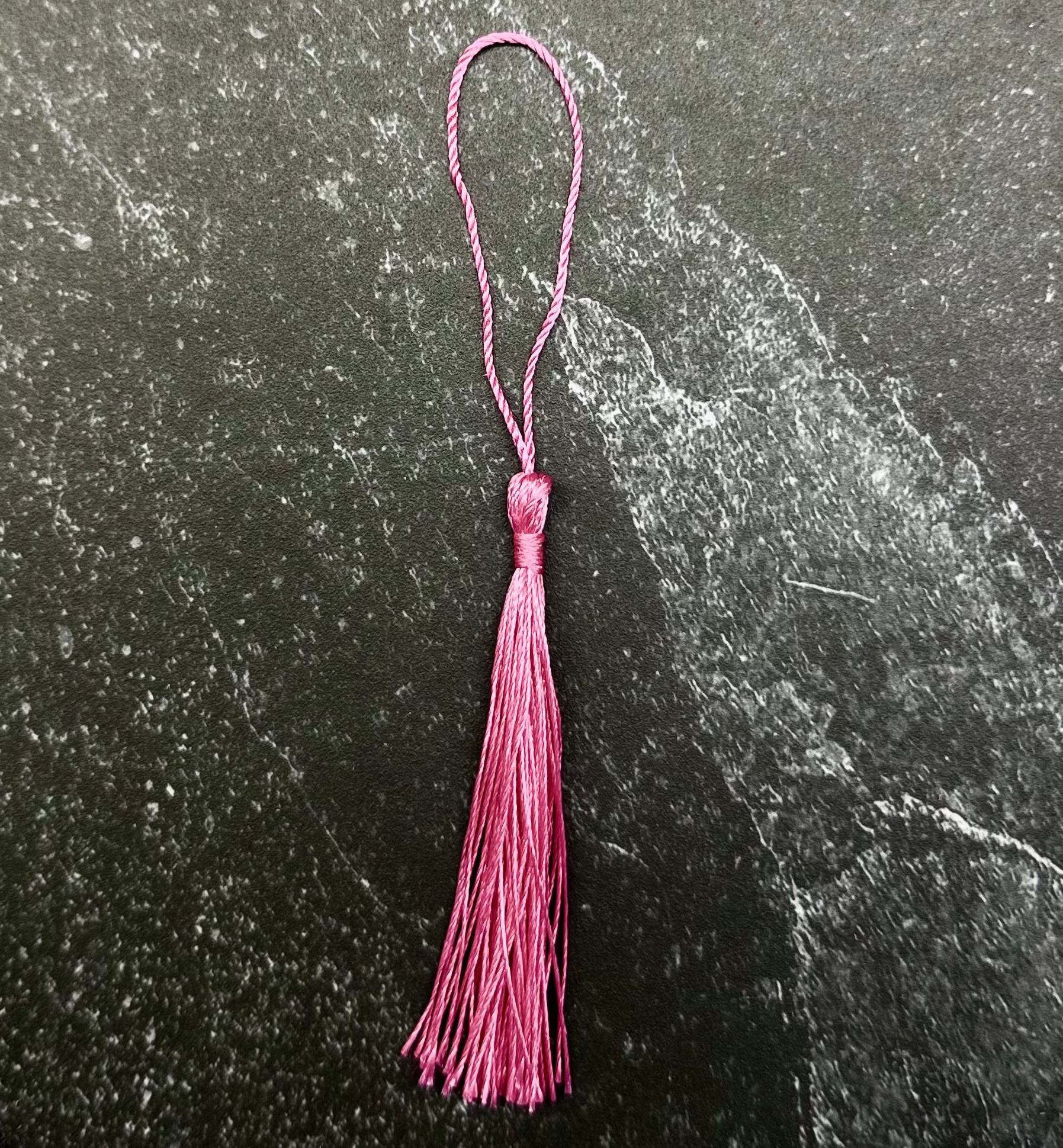 A pink tassel ornament with a hanging loop, displayed on a dark hexagonal stone tile against a black fabric background.