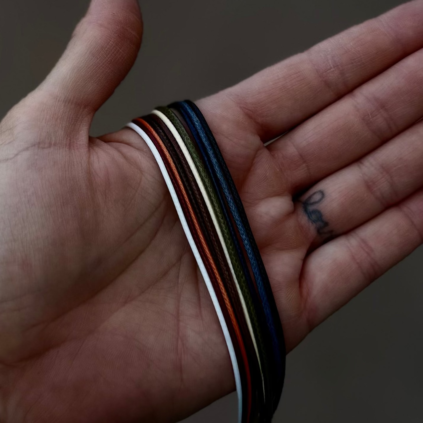 A close-up of a person's hand holding several thin multicolored wristbands, with a white smartwatch band on the wrist. The background is a blurred outdoor stone setting.