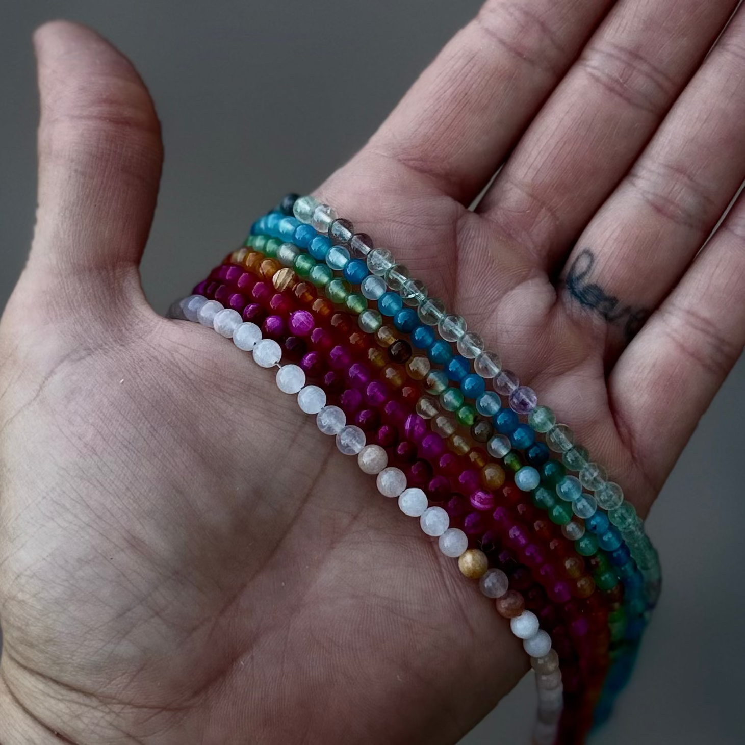 A close-up of a person's open hand holding a stack of colorful beaded bracelets, with a white smartwatch band and dark wristbands on the wrist, against a blurred gray background.