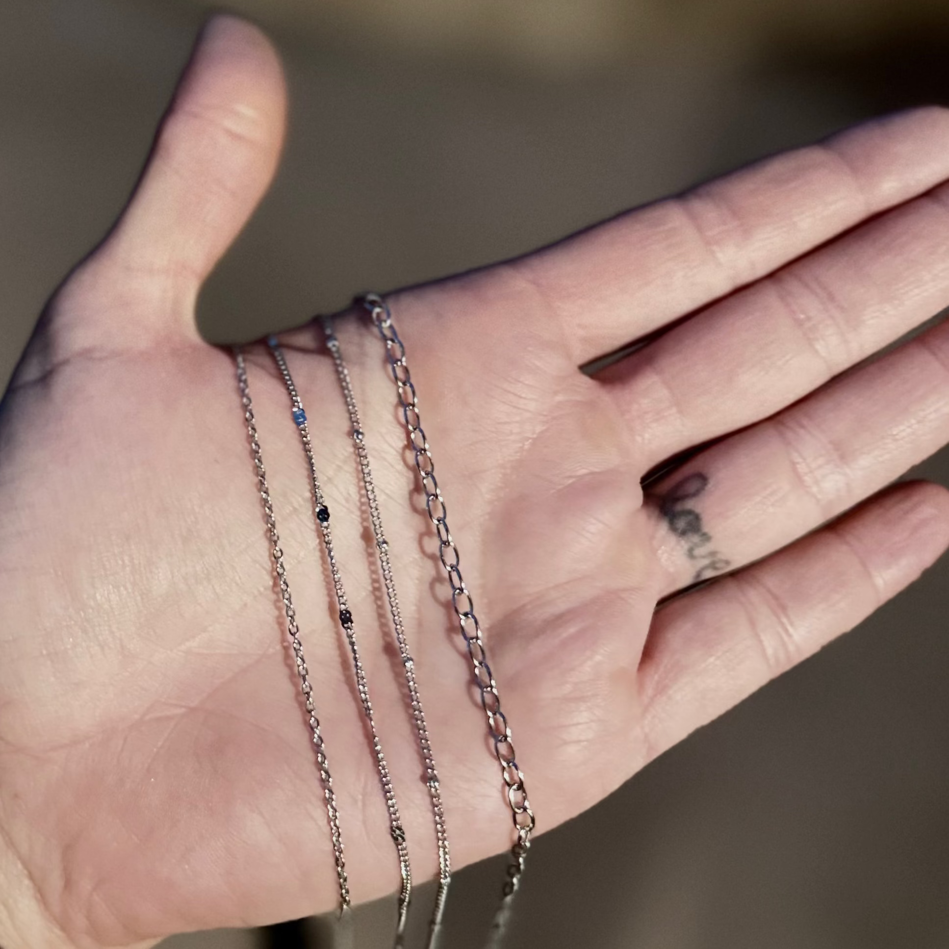 A close-up of a person's open palm holding three delicate rose gold chain necklaces, with a blurred outdoor background.