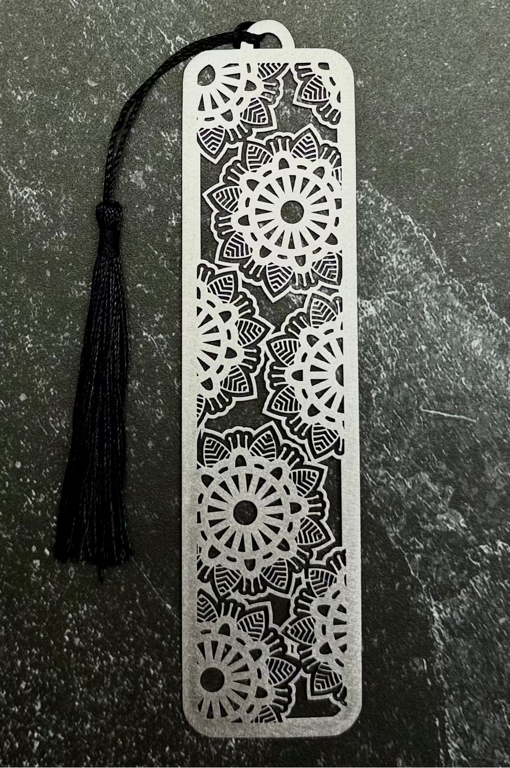 A silver metal bookmark with intricate floral cutout patterns and a black tassel, displayed on a dark green hexagonal stone slab against a dark navy fabric background.