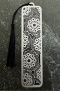 A silver metal bookmark with intricate floral cutout patterns and a black tassel, displayed on a dark green hexagonal stone slab against a dark navy fabric background.