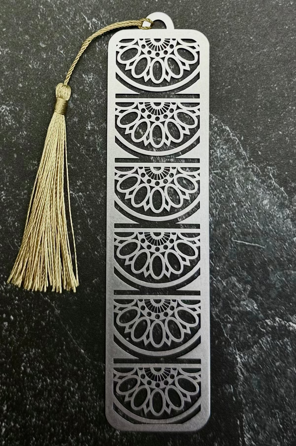 A silver metal bookmark with an intricate floral cutout pattern and a beige tassel, displayed on a dark gray hexagonal stone slab over a black fabric background.
