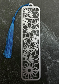 A silver metal bookmark with an intricate floral cutout pattern and a blue tassel, displayed on a dark gray hexagonal slate tile over a black fabric background.