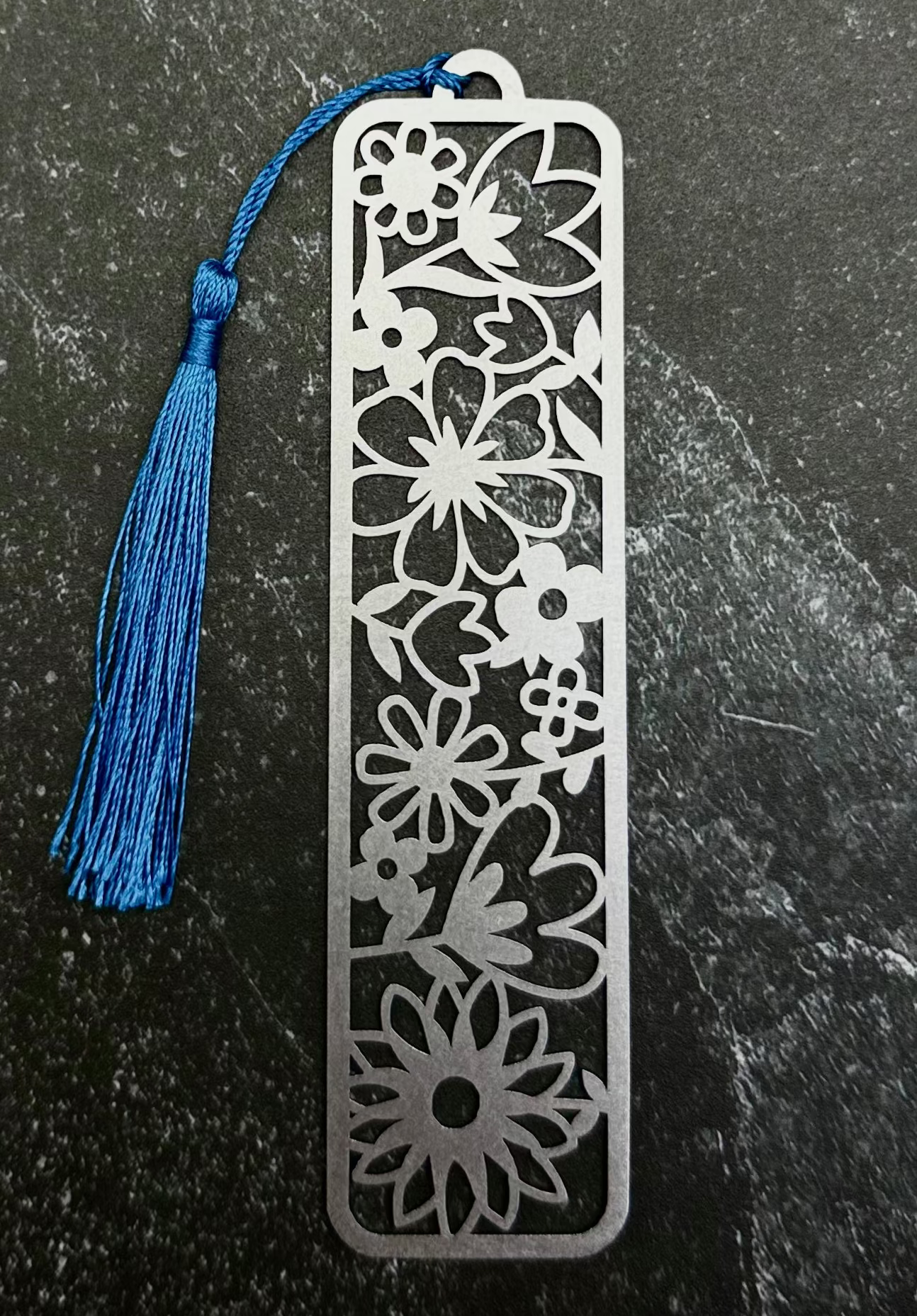 A silver metal bookmark with an intricate floral cutout pattern and a blue tassel, displayed on a dark gray hexagonal slate tile over a black fabric background.