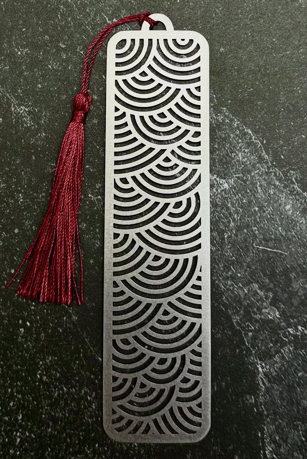 A silver metal bookmark with a red tassel, displayed on a dark green hexagonal stone slab over a black fabric background.