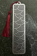 A silver metal bookmark with a red tassel, displayed on a dark green hexagonal stone slab over a black fabric background.