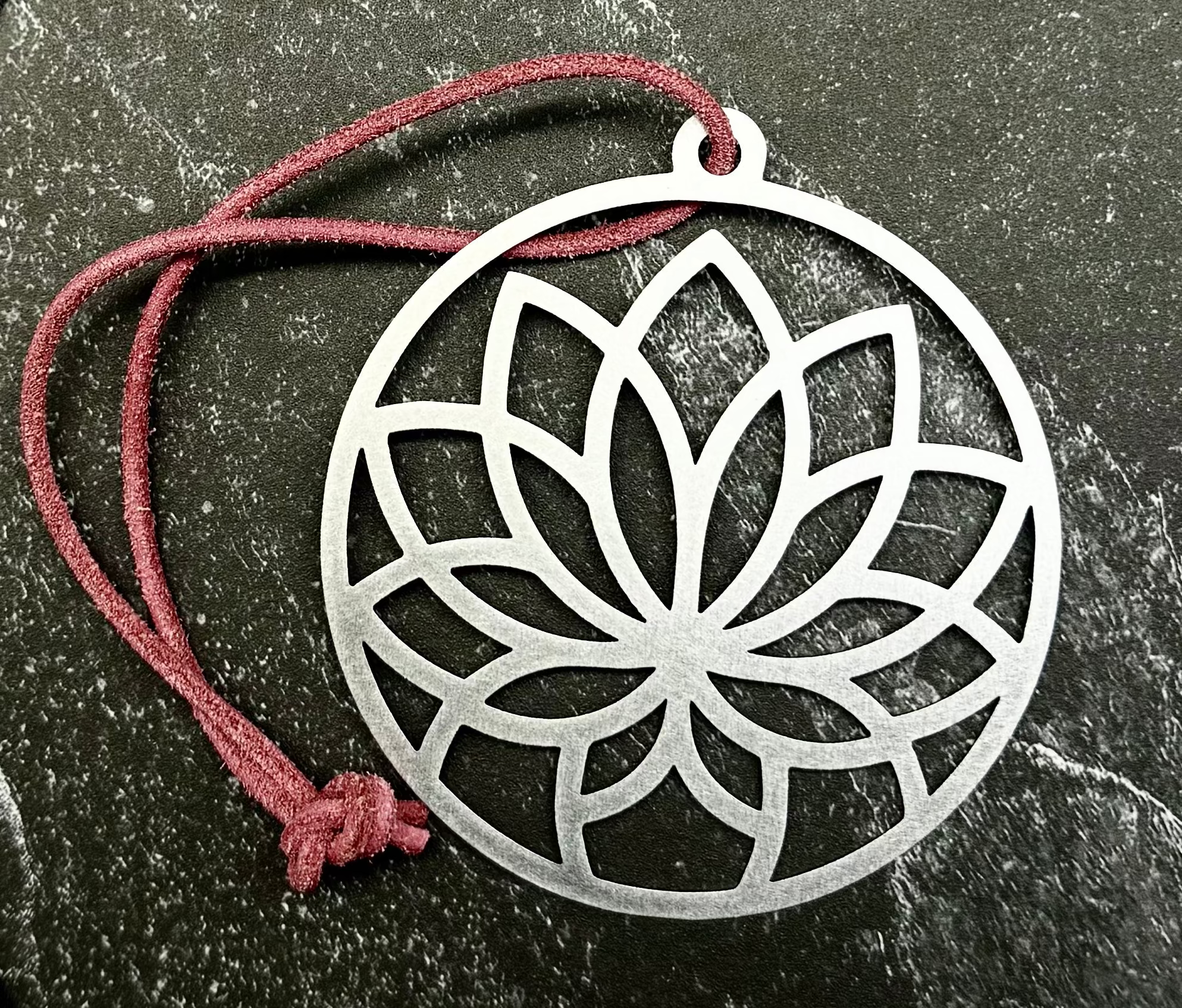 A silver metal lotus pendant with a red cord, displayed on a dark hexagonal stone slab over a black fabric background.