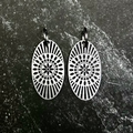 A pair of silver-toned circular dangle earrings with intricate sunburst patterns, displayed on a dark hexagonal stone slab set against a black fabric background.