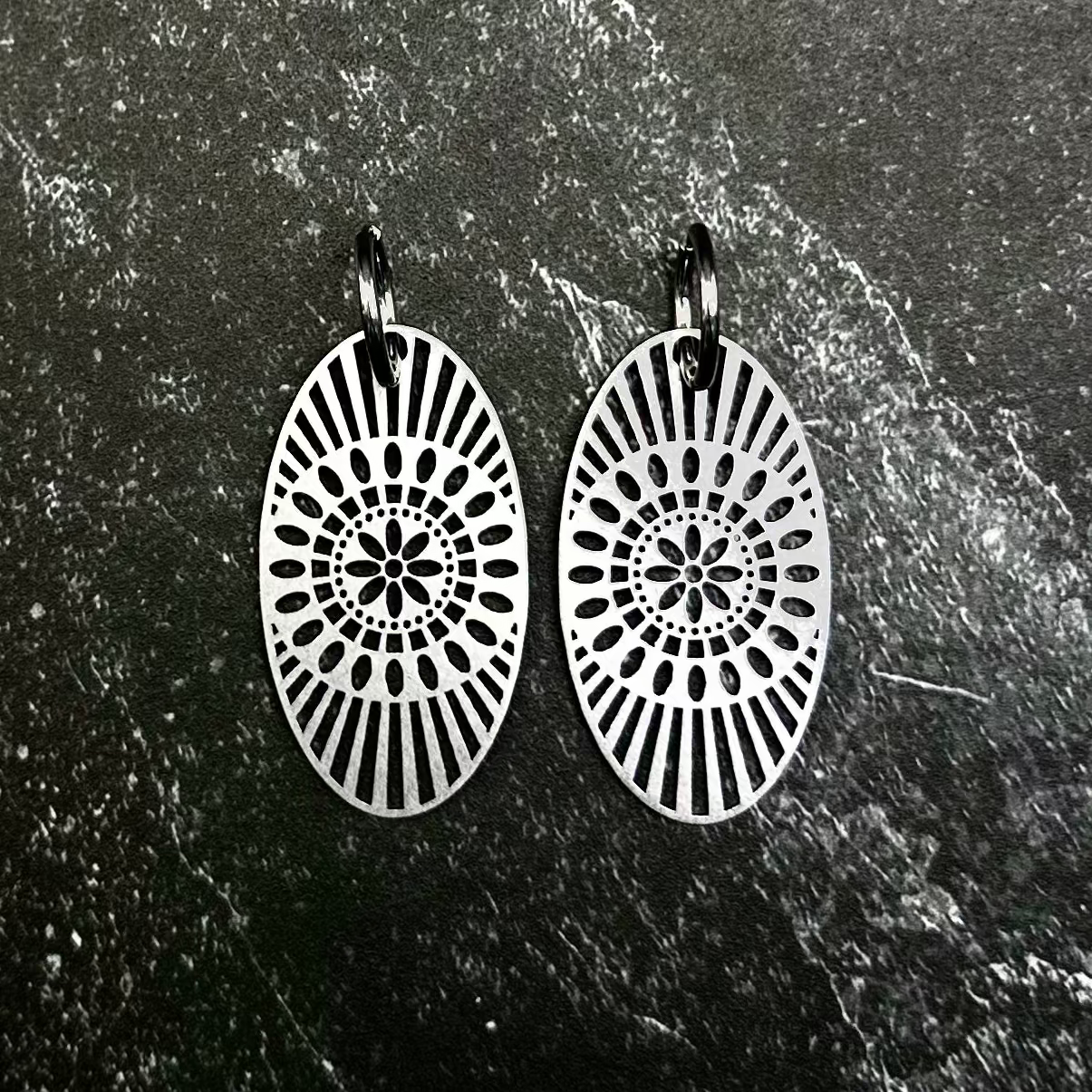 A pair of silver-toned circular dangle earrings with intricate sunburst patterns, displayed on a dark hexagonal stone slab set against a black fabric background.