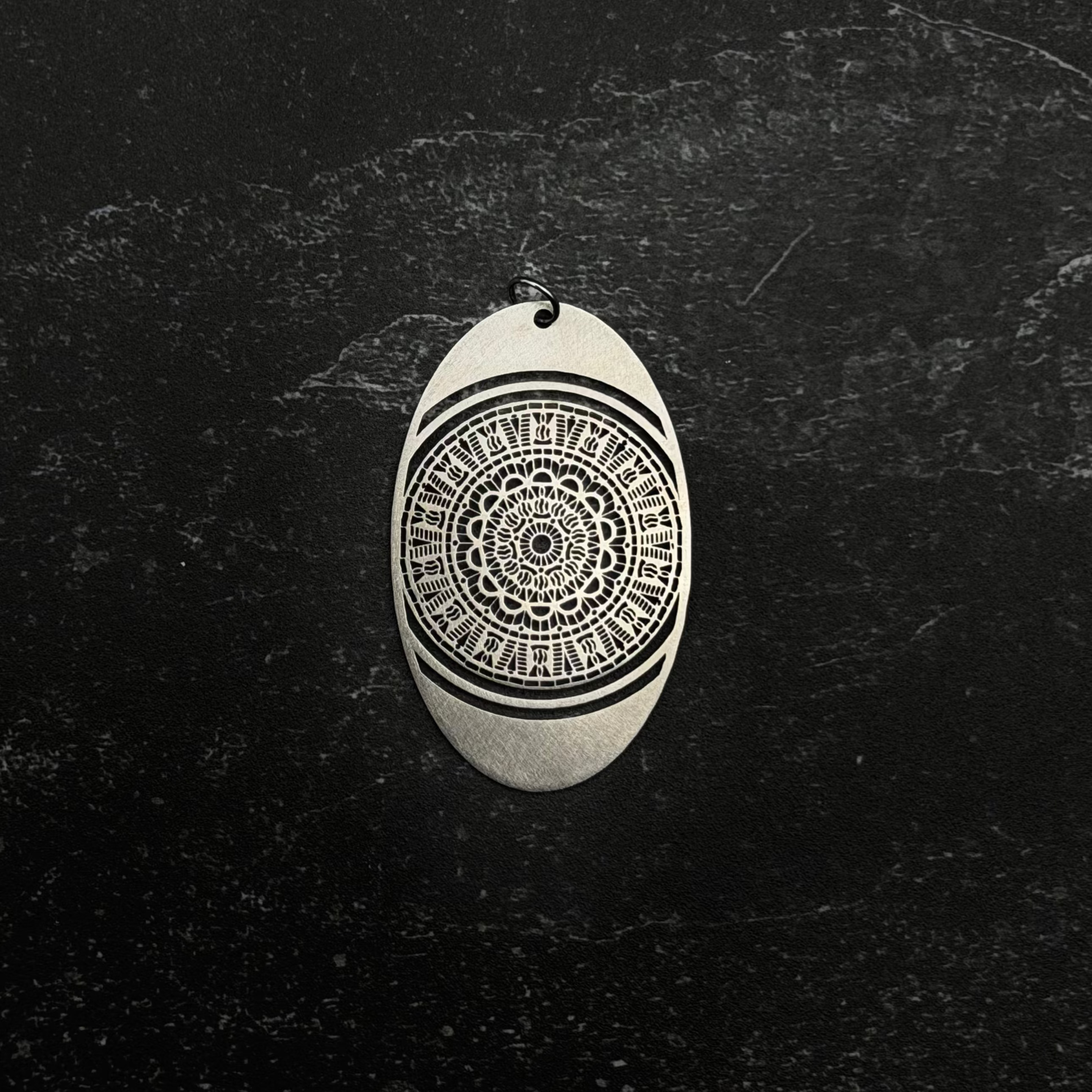 Stainless steel Oval-shaped pendant with intricate black pattern on a dark textured background