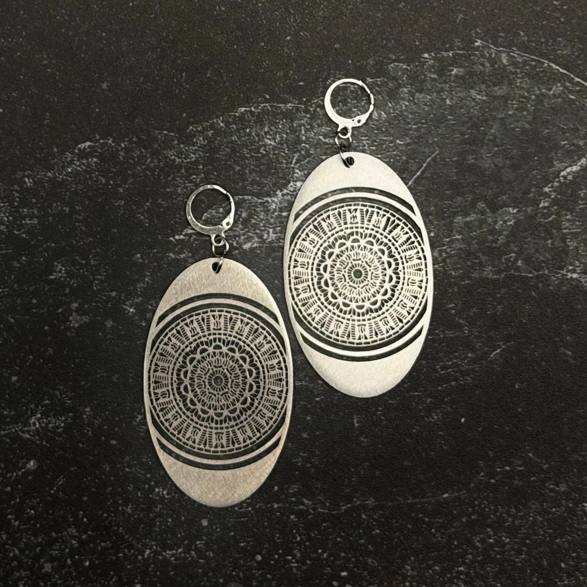 Stainless steel oval earrings with mandala design on a dark textured surface