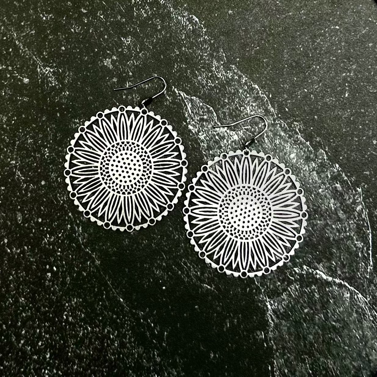 A close-up of a black textured fabric garment with a hexagonal patch showing two silver-toned circular mandala earrings on a dark green stone background.