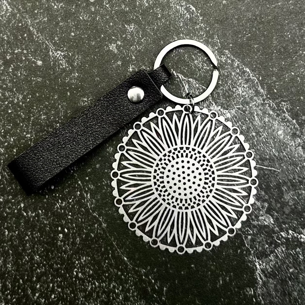 A silver-toned circular keychain with a black leather loop and keyring, displayed on a dark hexagonal stone slab over a black fabric background.
