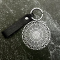 A silver-toned circular keychain with a black leather loop and keyring, displayed on a dark hexagonal stone slab over a black fabric background.
