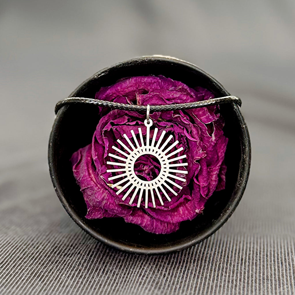 Silver sun-shaped pendant on a black cord with a purple flower in a black dish on a gray background