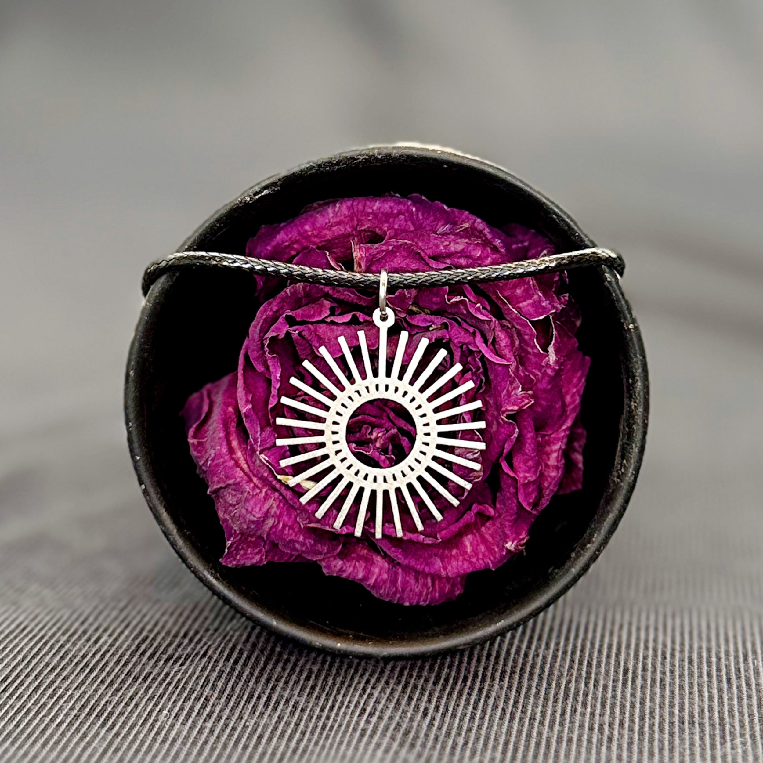 Silver sun-shaped pendant on a black cord with a purple flower in a black dish on a gray background